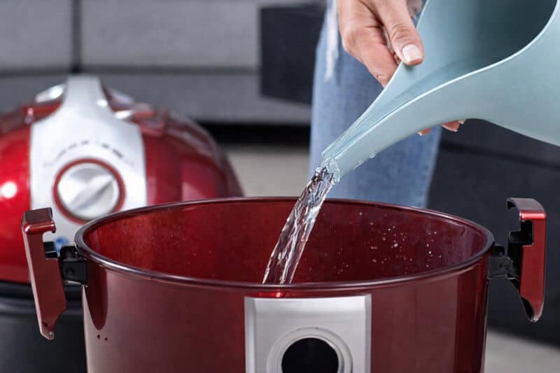 Person pouring clean water into the transparent water chamber of a water filtration vacuum cleaner, demonstrating how the vacuum’s water-based filtration system is filled before use.