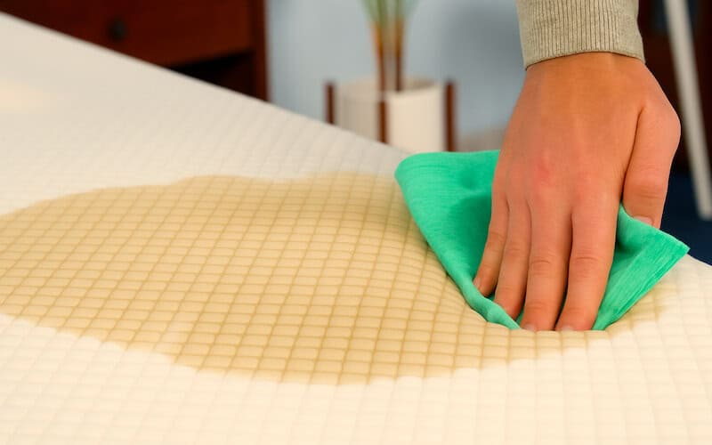 A hand using a green microfiber cloth to blot a light brown stain on a white, textured mattress surface.