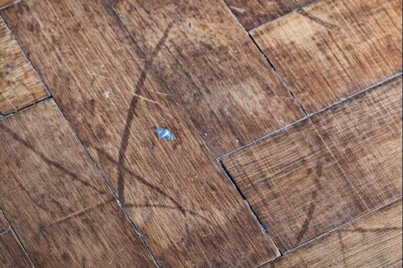 Damaged parquet wood flooring showing surface scratches, faded finish, small gaps between blocks, and signs of wear.